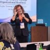 Ashley, wearing a black blazer and an orange lanyard with her name badge, holds a microphone and adjusts her glasses while speaking passionately in front of a projection screen. The session focuses on people, training, structure, and process, as indicated by the slide. In the foreground, an audience member with gray hair, dressed in a floral blouse, listens attentively. The setting includes a wooden podium, water bottles on the table, and a modern conference room with a teal wall and a small monitor displaying the Clay logo. This photo, part of the event's photo gallery on Utilityevents.com, highlights the educational and collaborative spirit of the conference.