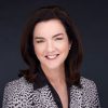 A professional headshot of Barbara Trautlein smiling, wearing a patterned blazer and black blouse against a dark background.